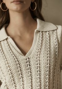 Pull tricoté en beige clair avec encolure en V et col, présentant un motif en points ouverts texturé et des manches courtes.
