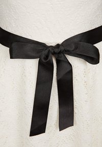 Biała koronkowa sukienka z grubą, teksturowaną czarną satynową wstążką związaną w kokardę w talii, eksponująca skomplikowane detale koronkowe oraz gładką tkaninę.