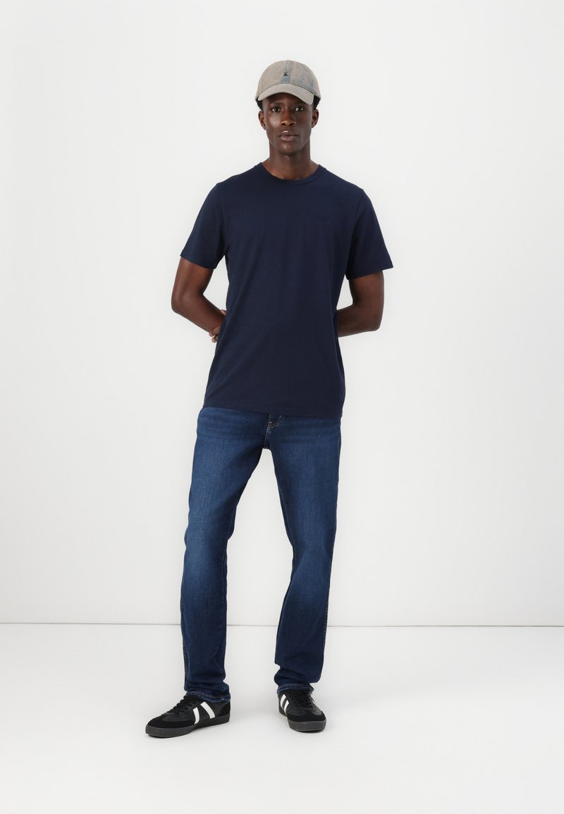Camiseta azul marino, jeans azul, zapatillas negras con acentos blancos y una gorra gris claro. El modelo está de pie frente a un fondo blanco liso.