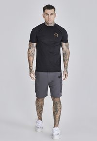 Schwarzes Kurzarm-Sportshirt aus strukturiertem Stoff mit goldenem Logo. Kombiniert mit grauen Cargo-Shorts mit Seitentaschen. Weiße Turnschuhe.