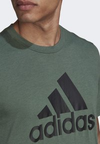 Grön t-shirt med kort ärm i mjukt tyg som har en stor svart adidas-logotyp över bröstet, rund halsringning och ribbade kanter.