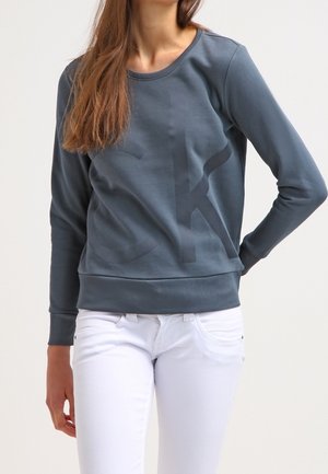 Kobieta w długim ciemnoszarym swetrze z subtelnym dużym logo i białych dopasowanych jeansach, stojąca na jasnym tle.