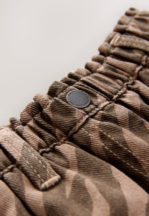 Brunt tekstureret stof med et camouflagemønster, der har rynkede detaljer og en metal knap med præget tekst på et stykke tøj.