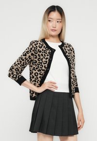 Kvinne iført en beige og svart leoaprdprint cardigan over en hvit topp, kombinert med en mørkegrå plisserte skjørt, stående med den ene hånden på hoften.