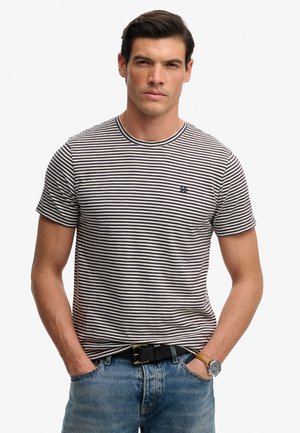 Mann trägt ein schwarz-weiß gestreiftes T-Shirt, blaue Jeans, schwarzen Gürtel und Armbanduhr, steht mit den Händen in den Taschen vor neutralem Hintergrund.