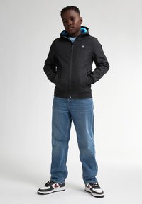 Schwarze Zip-Per-Hoodie mit blauem Innenfutter, mit einem Logo auf der Brust. Kombiniert mit blauen Jeans und Schwarz/Weißen Sneakern.