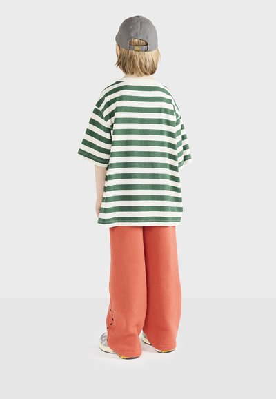 Stribet t-shirt i grøn og hvid, parret med orange, løstsiddende bukser. En grå kasket bæres, og klodsede sneakers er synlige.