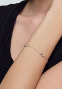 Srebrna bransoletka łańcuszkowa z okrągłymi ogniwami i teksturowanym akcentem. Noszona na nadgarstku, ukazuje gładką skórę i ciemny sweter.