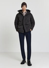 Veste matelassée noire avec capuche, deux poches avant, portée sur une chemise claire. Associée à un pantalon bleu marine et des bottines noires. Bonnet en laine marron.