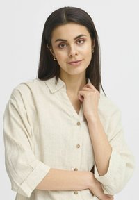 Giovane donna con lunghi capelli scuri, che indossa una camicia beige con bottoni e piccoli orecchini a cerchio dorati, in posa con una mano vicino al mento.