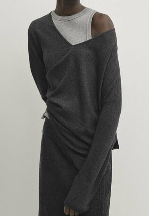 Personne portant un pull gris foncé à épaules dénudées sur un haut sans manches gris clair, sur un fond clair uni.