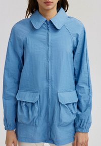 Jachetă ușoară, de culoare albastră deschisă, cu guler, fermoar frontal și două buzunare mari tip patch, având accente din material pliat.