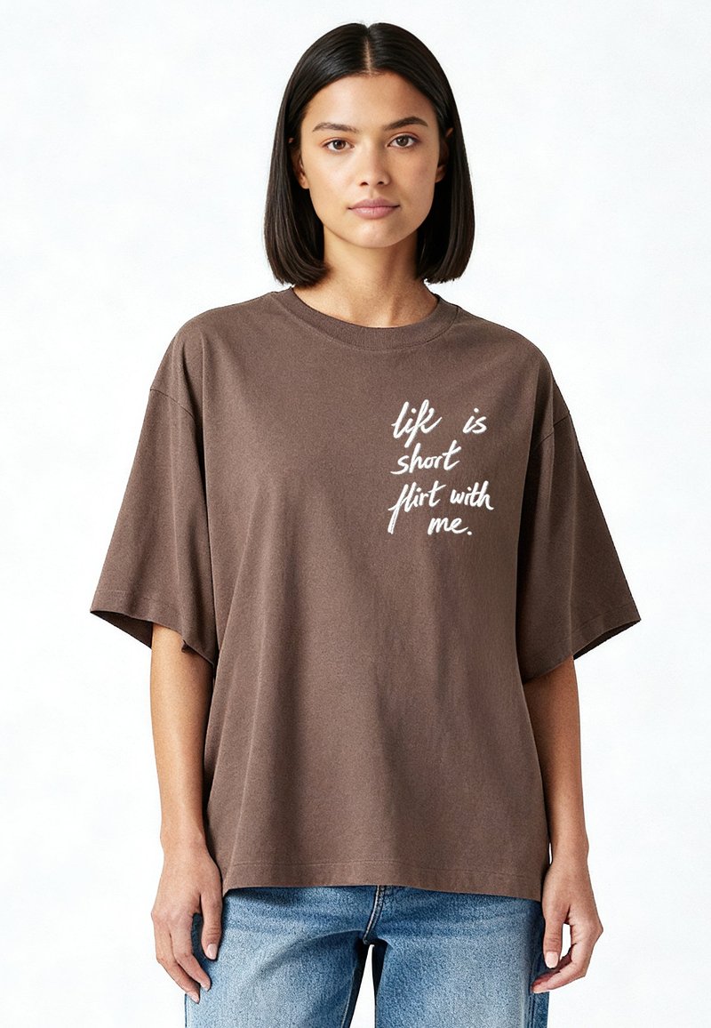 Jeune femme aux cheveux foncés et raides portant un t-shirt marron oversize avec un texte en script blanc et un jean bleu, debout devant un fond clair.