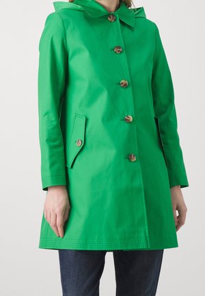 Femme portant un manteau vert vif boutonné avec des poches à rabat et un col, associé à un jean bleu foncé sur un fond uni.