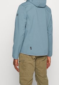 Giacca softshell blu con cappuccio, caratterizzata da una texture liscia, con un logo sulla manica e un'etichetta vicino all'orlo, abbinata a pantaloni cargo color khaki.