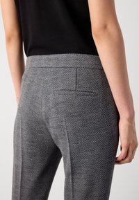 Personne portant un pantalon gris à taille haute en chevron avec une seule poche passepoilée à l’arrière et un haut noir sans manches sur un fond uni.
