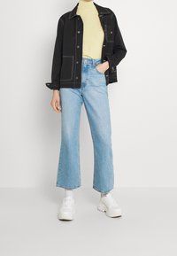 Schwarze Denimjacke mit weißen Nähten, gelb gepunkteter Rollkragenpullover, hellblaue weit geschnittene Jeans und weiße klobige Sneakers.