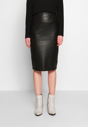 Kobieta ubrana w czarną, skórzaną spódnicę ołówkową, czarną bluzkę z długim rękawem oraz białe botki w wzór skóry węża, stojąca na tle gładkiego tła.