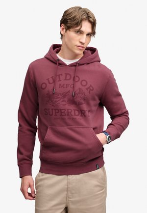 Młody mężczyzna w bordowej bluzie z kapturem z napisem "Outdoor Superdry" i grafiką góry, beżowych spodniach i zegarku na nadgarstku, pozujący z jedną ręką w kieszeni.