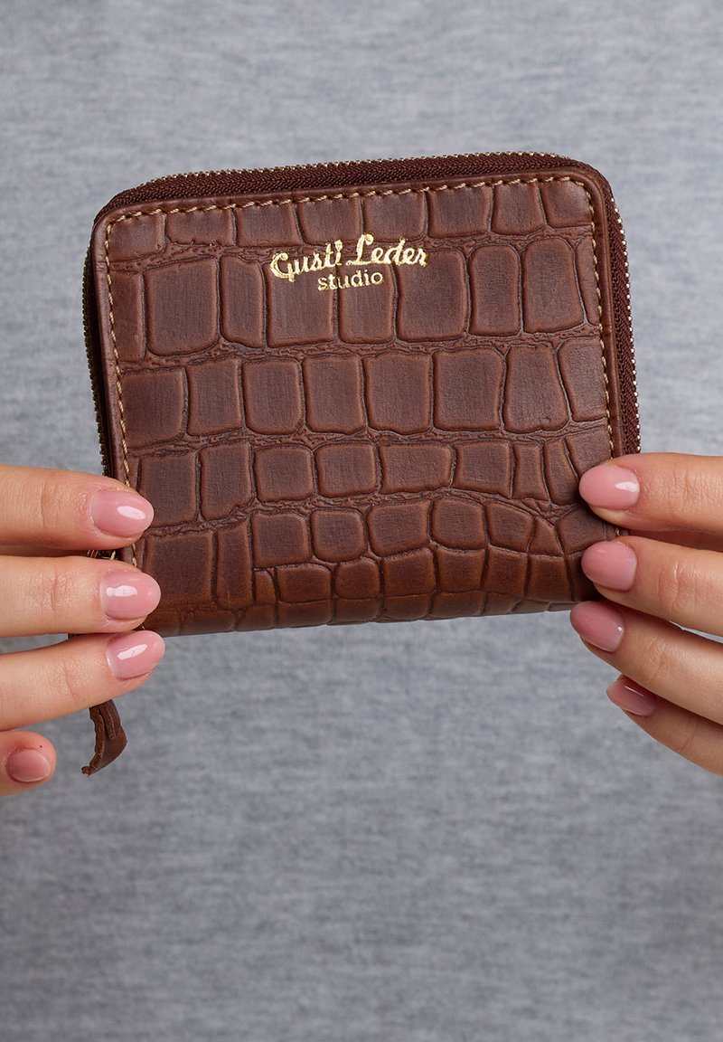 Hnědá peněženka z krokodýlí kůže s embosovaným zlatým logem, na zip a texturovaným povrchem, držená mezi rukama s růžovým nalakováním nehtů.