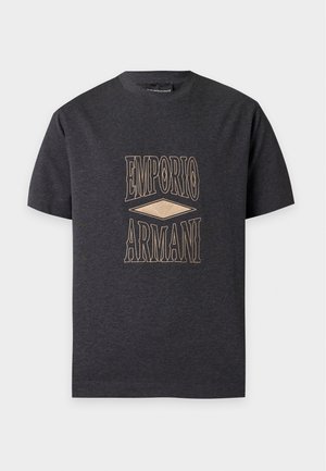 Jeune homme portant un t-shirt Emporio Armani gris foncé, un jean large bleu clair et des baskets blanches, debout avec une main dans la poche.