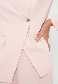 Κοντινό πλάνο χεριού στην τσέπη σακακιού ανοιχτό ροζ κοστουμιού, κλεισμένο με χρυσό κουμπί στροβίλου.