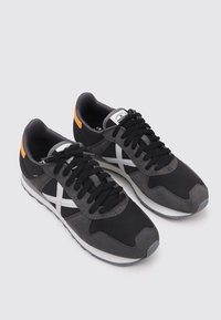 Zapatillas negro y gris con un upper de ante y malla, diseño cruzado en blanco, agujetas negras, suela gris y un acento naranja en el talón.