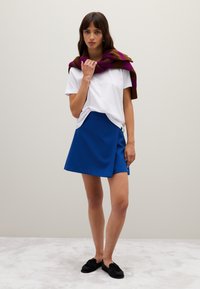 Frau steht da, trägt ein weißes T-Shirt, einen blauen Rock, schwarze Slipper und einen braun-lila gestreiften Pullover über den Schultern.