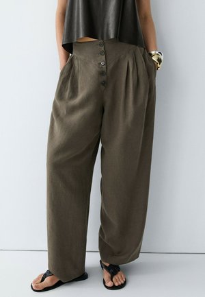 Pantalon marron ample taille haute avec boutonnière devant, porté avec un top court noir, des sandales noires et un large bracelet doré et argenté.