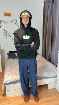 Noor mees musta kapuutsiga ja beežipeaga seismas ristatud kätega magamistoas, kus on voodi, hallid kardinad ja seinakaunistused.