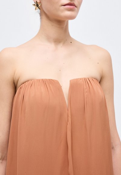 Kvinde iført stropløs, ferskenfarvet kjole med plisserede bodicedetaljer og stor guldfarvet stjerneformet ørering i venstre øre.