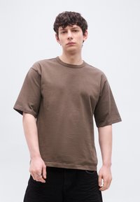 Νεαρός άνδρας με σγουρά σκούρα μαλλιά, φορώντας ένα καφέ φαρδύ μπλουζάκι και μαύρο παντελόνι, στέκεται μπροστά σε ένα απλό ανοιχτόχρωμο φόντο.