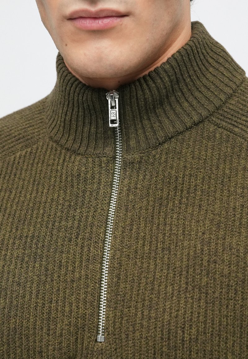 Oliven grøn ribbet sweater med høj krave og sølv metal lynlås. Stof virker struktureret og tykt, velegnet til koldt vejr.