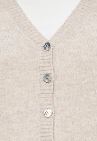 Beige kötött kardigán V-nyakkal, három fém kagylómintás gombbal és bordás textúrával az éleken.