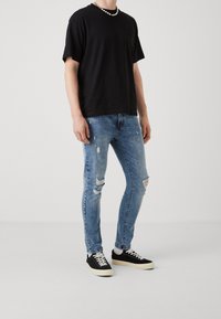 Camiseta negra de manga corta con bolsillo; vaqueros desgastados de mezclilla azul claro con roturas; zapatillas negras con suelas blancas.