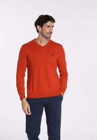 Pull rouge à col en V fabriqué dans un matériau tricoté lisse, doté de manches longues et d'un petit logo bleu sur la poitrine. Porté avec un pantalon marine.