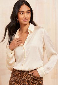 Bluză satin crem cu guler cu nasturi, mâneci lungi și finisaj texturat, asociată cu pantaloni din material cu imprimeu leopard maro.