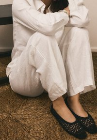 Ballerines noires en crochet avec un motif aéré. Portées avec un pantalon ample en lin blanc. Tapis en fibres naturelles texturé en dessous.