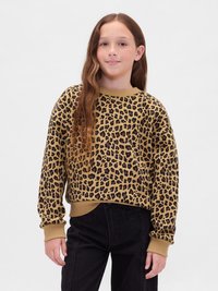 Ragazzina con lunghi capelli mossi, indossa un pullover leopardato beige e nero e pantaloni neri, in piedi su uno sfondo bianco semplice.