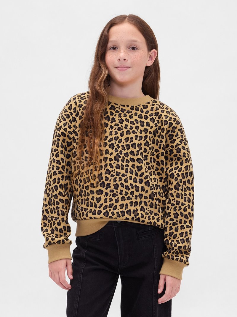 Ragazzina con lunghi capelli mossi, indossa un pullover leopardato beige e nero e pantaloni neri, in piedi su uno sfondo bianco semplice.
