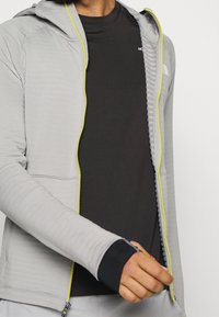 Sudadera de cremallera gris claro con cremallera amarilla de contraste, textura acanalada y orificios para los pulgares. Llevada sobre una camiseta negra de manga corta.