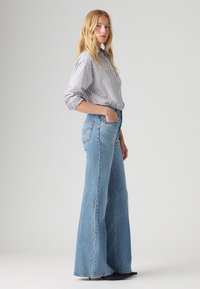 Jean flare bleu clair à taille haute avec un design classique à cinq poches, associé à une chemise à rayures à boutons, montrée de profil.