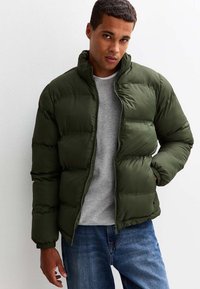 Grön puffjacka med hög krage, dragkedjestängning och quiltat mönster. Bärs över en grå sweatshirt, ihop med blå jeans.