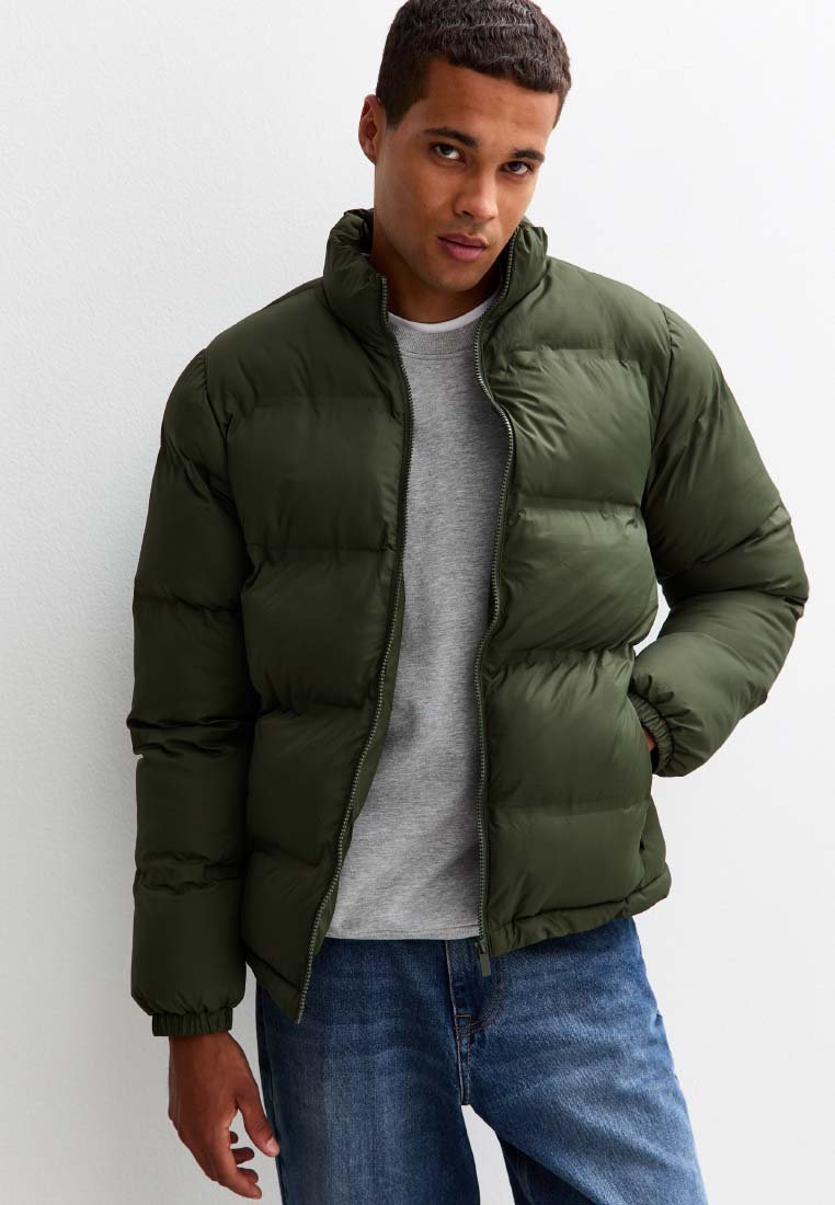 Grön puffjacka med hög krage, dragkedjestängning och quiltat mönster. Bärs över en grå sweatshirt, ihop med blå jeans.