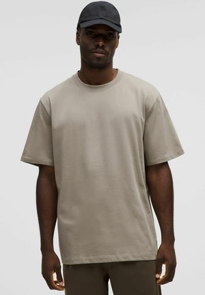Mann in einem beigen, übergroßen T-Shirt mit kurzen Ärmeln und dunklen Hosen, der mit einer schwarzen Baseballkappe vor einem schlichten hellen Hintergrund steht.