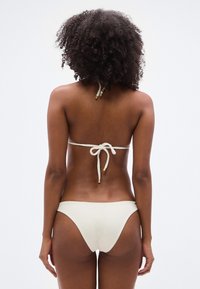 Διπλό μπικίνι σε ανοιχτό κρεμ χρώμα, με υφασμάτινη υφή ριμπ, ρυθμιζόμενους δερούς και ελάχιστη κάλυψη στο κάτω μέρος.
