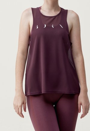 Mujer con camiseta deportiva sin mangas color granate y leggings a juego, de pie contra un fondo claro y liso.