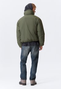 Olivgrüne Steppjacke mit hohem Kragen, getragen über einem dunklen Oberteil und blauer Jeans. Die Jacke hat eine glatte Textur und eine lockere Passform.