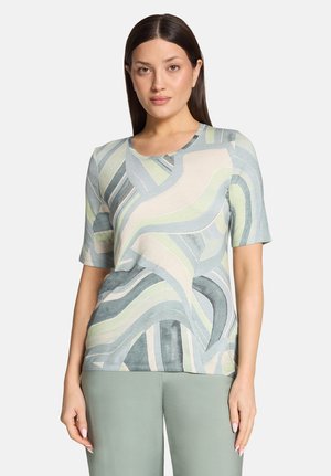 Frau mit glatten dunklen Haaren, die ein kurzärmeliges Oberteil mit pastellfarbenem abstraktem Muster und hellgrüne Hosen trägt, steht vor einem einfarbigen Hintergrund.