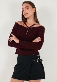 Sieviete, kura valkā bordo off-shoulder ribota džemperi ar sirds pogām un melnu svārku ar sudraba sprādžu detaļām, rokas savilktas.